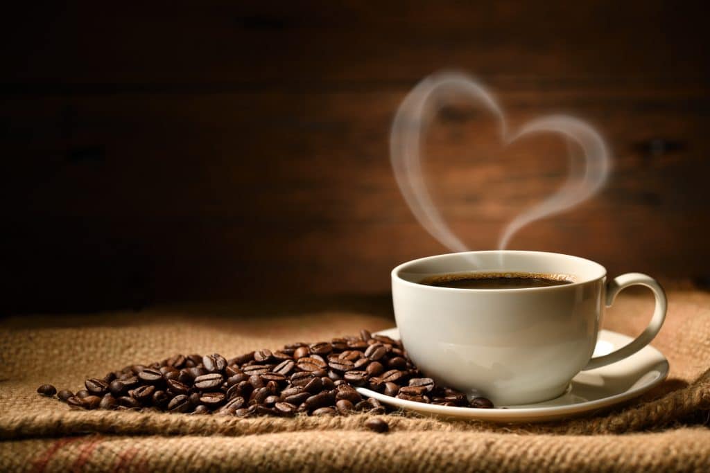 tasse de café chaud avec coeur en fumée posé sur une toile de jute avec des grains de café