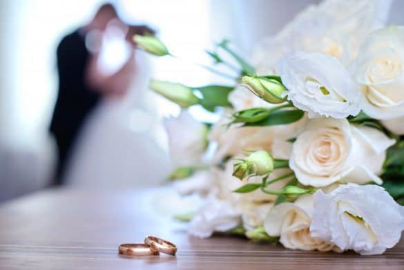 alliances posées sur une table à côté d'un bouquet de fleurs et couple en fond